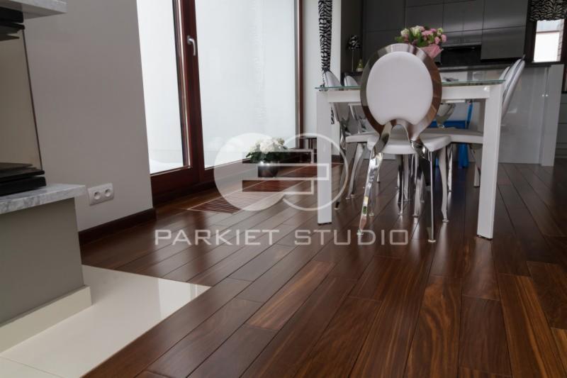 Ciemnobrązowa podłoga z egzotycznego drewna ułożona w jadalni, widoczny fragment ściany z listwą przypodłogową i białą płytką, stół z przezroczystym blatem i białymi krzesłami w tle.