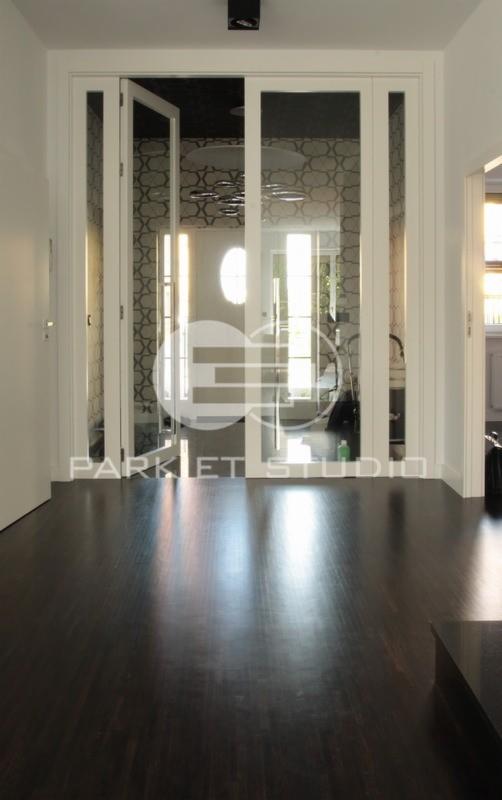 Ciemna, lakierowana podłoga drewniana z widocznymi refleksami światła, prowadząca do białych, przeszklonych drzwi wewnętrznych z jednym skrzydłem otwartym, za którymi widać wzorzystą tapetę...