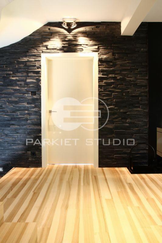 Jasna podłoga z parkietu ułożona w pasy, białe drzwi wewnętrzne z klamką, ściana wyłożona ciemnym kamieniem dekoracyjnym podświetlona lampą sufitową.