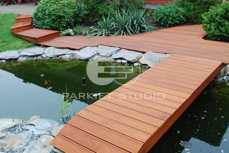 Drewniany taras z pomostem nad oczkiem wodnym, schodki prowadzące z trawnika, roślinność ogrodowa w tle, widoczne deski tarasowe z bliska.