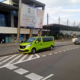 Jaskrawo zielony bus Renault Trafic parkuje przed nowoczesnym budynkiem lotniska z dużym billboardem reklamowym. Na pierwszym planie pasy dla pieszych.