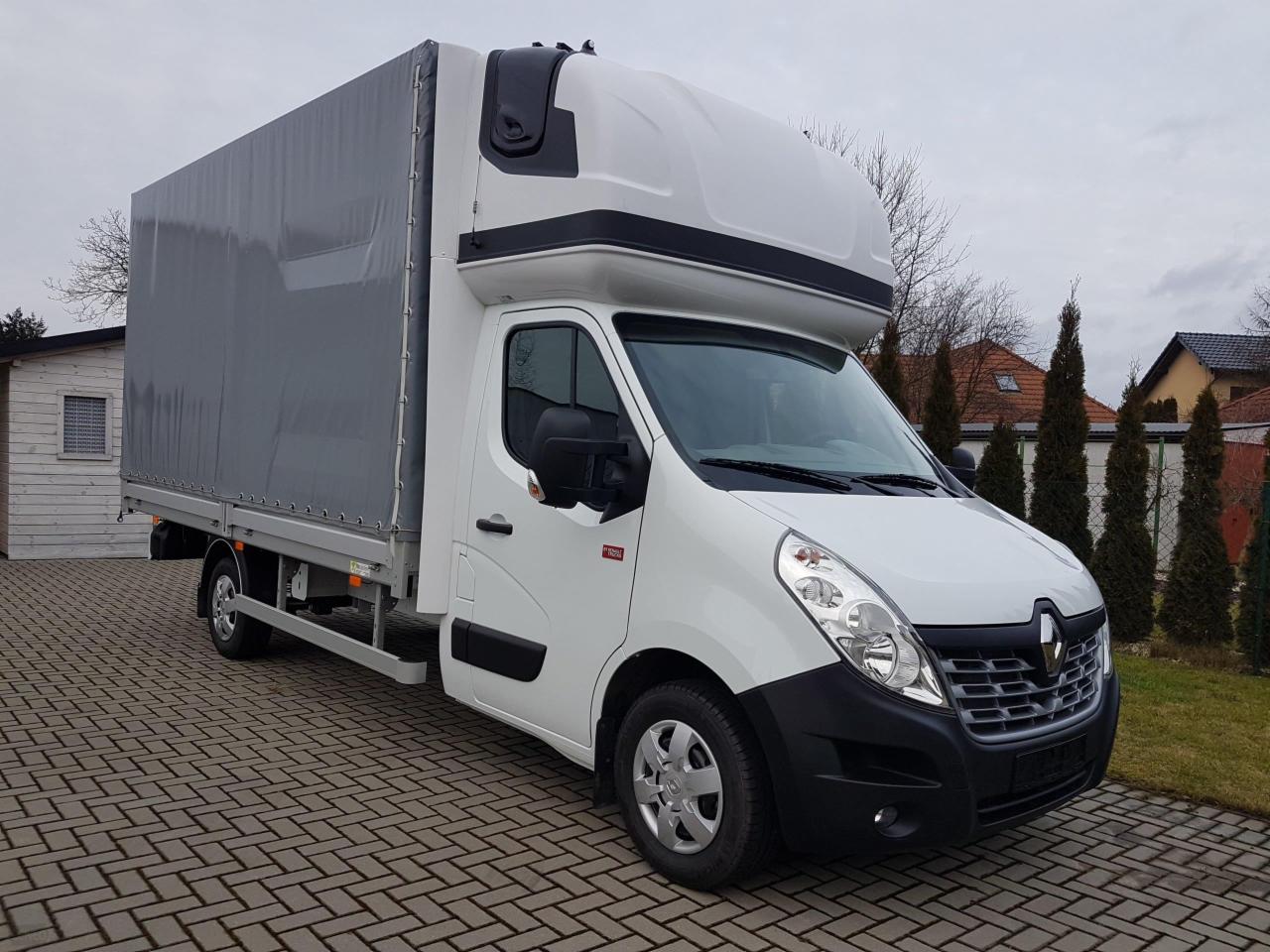 Biały samochód dostawczy Renault Master z zabudową kontenerową i szarą plandeką, zaparkowany na brukowanej nawierzchni, z budynkami mieszkalnymi i zielenią w tle.