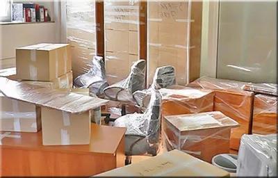Spakowane biuro: kartony, meble biurowe owinięte folią, przygotowane do transportu. Widoczne biurka, krzesła obrotowe i regały zabezpieczone przed uszkodzeniem.