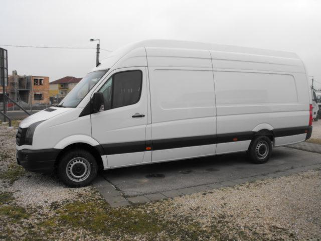 Biały, wysoki furgon dostawczy marki Volkswagen Crafter, zaparkowany na betonowej nawierzchni z widocznym placem budowy w tle, sfotografowany z boku w pochmurny dzień.