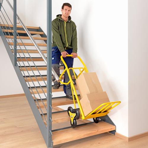 Mężczyzna w zielonej bluzie i roboczych spodniach wciąga po schodach w domu żółty wózek z dwoma kartonami.