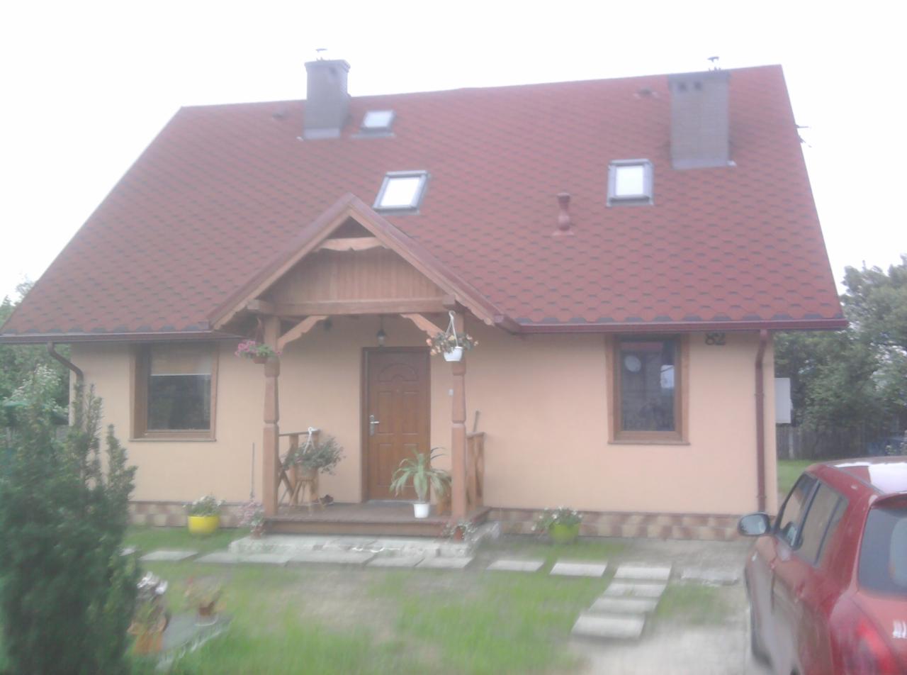 Dom szkieletowy w stanie surowym zamkniętym: widok na elewację frontową z drewnianym gankiem, czerwonym dachem z lukarnami i kominem, oraz częściowo widocznym samochodem osobowym.