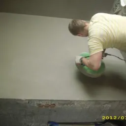 Pracownik w koszulce w paski szlifuje ścianę szlifierką oscylacyjną z zieloną tarczą, widoczny fragment ściany po obróbce i surowy beton u dołu.