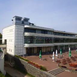 Widok z góry na dwupiętrowy budynek trybuny z przeszkloną fasadą, z flagami różnych państw na balkonie, z zewnętrznym tarasem z parasolami i ławkami.