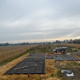 E&D HOUSE SPÓŁKA Z OGRANICZONĄ ODPOWIEDZIALNOŚCIĄ - Zbrojenie fundamentów domu jednorodzinnego na tle pola kukurydzy. Widoczne szalunki, pręty zbrojeniowe, folia budowlana i ekipa budowlana przy barakowozie.