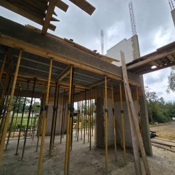 E&D HOUSE SPÓŁKA Z OGRANICZONĄ ODPOWIEDZIALNOŚCIĄ - Wnętrze budynku w stanie surowym otwartym. Żółte stemple podtrzymują strop, widoczne ściany z betonu komórkowego i wystające zbrojenia. Na zewnątrz zieleń i samochód.