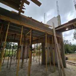 Wnętrze budynku w stanie surowym otwartym. Żółte stemple podtrzymują strop, widoczne ściany z betonu komórkowego i wystające zbrojenia. Na zewnątrz zieleń i samochód.