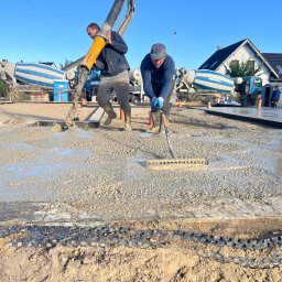 E&D HOUSE SPÓŁKA Z OGRANICZONĄ ODPOWIEDZIALNOŚCIĄ - Wylewanie betonu na przygotowany teren przez trzech robotników. W tle betoniarki i dom jednorodzinny z dwuspadowym dachem w słoneczny dzień.