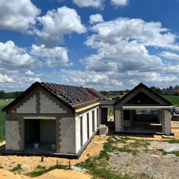 E&D HOUSE SPÓŁKA Z OGRANICZONĄ ODPOWIEDZIALNOŚCIĄ - Dom z garażem w stanie surowym otwartym, z jasnymi ścianami z bloczków i częściowo ułożonymi ciemnymi dachówkami, pod błękitnym niebem z chmurami.