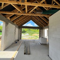 E&D HOUSE SPÓŁKA Z OGRANICZONĄ ODPOWIEDZIALNOŚCIĄ - Wnętrze budowanego domu z widoczną drewnianą konstrukcją dachu, białymi ścianami z bloczków, betonową posadzką i zielonymi przewodami wentylacyjnymi. W tle zieleń.
