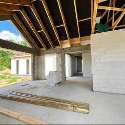 E&D HOUSE SPÓŁKA Z OGRANICZONĄ ODPOWIEDZIALNOŚCIĄ - Wnętrze budowanego domu z widoczną drewnianą konstrukcją dachu, białymi ścianami z bloczków i betonową posadzką. Na pierwszym planie leżą drewniane belki. W tle zielony krajobraz i błękitne niebo.