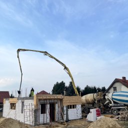 E&D HOUSE SPÓŁKA Z OGRANICZONĄ ODPOWIEDZIALNOŚCIĄ - Budowa domu z białych bloczków, wylewanie stropu betonem z pompy. Żółte ramię pompy i niebiesko-biała betoniarka na tle częściowo zachmurzonego nieba.