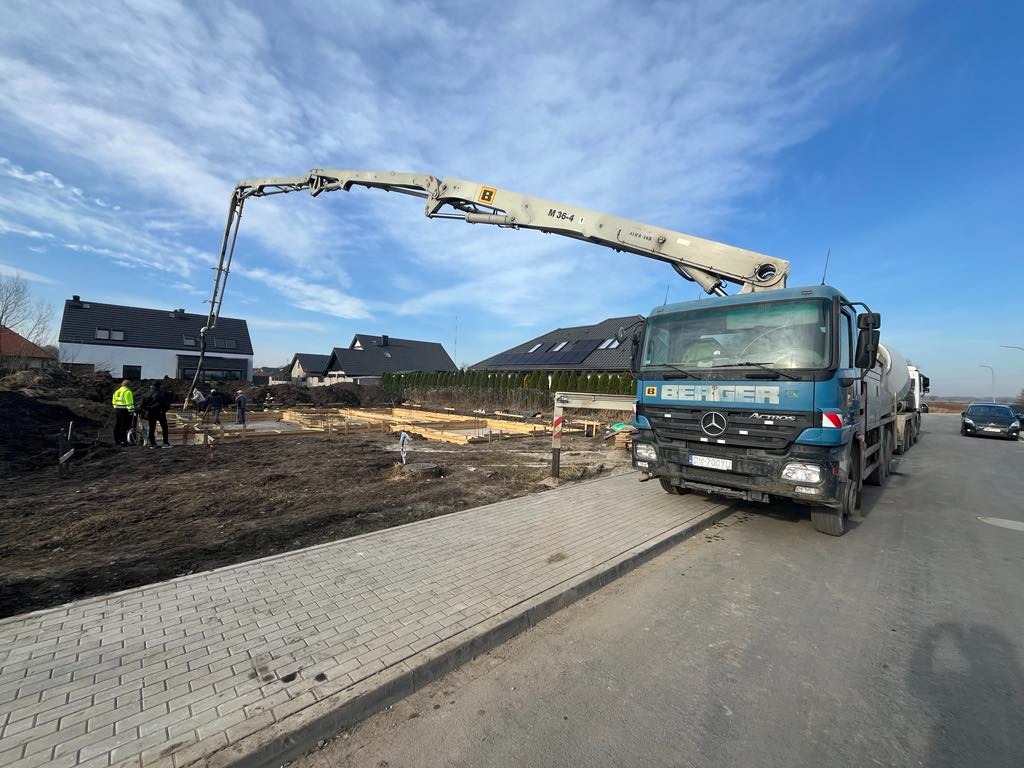 Żurawina: Betonowanie fundamentów domu jednorodzinnego z użyciem pompy do betonu marki Berger. Widoczny szalunek i pracownicy w odblaskowych kamizelkach.