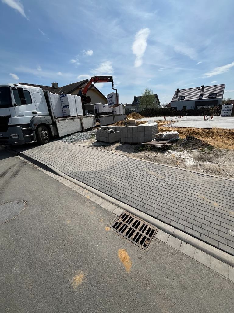 Ciężarówka z dźwigiem dostarcza bloczki Solbet na plac budowy domu jednorodzinnego. Widoczne fundamenty i podjazd z kostki brukowej. Żórawina.