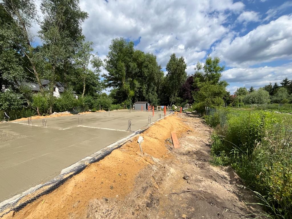 Wylana płyta fundamentowa z wystającymi prętami zbrojeniowymi na działce w Jarnołtowie, Wrocław. Widok na teren budowy w słoneczny dzień.