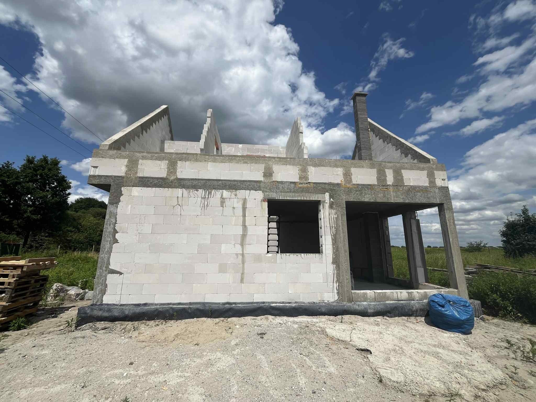 Niedokończony budynek z białych bloczków, widoczny komin i otwory okienne. W tle niebo z chmurami i zieleń. Na pierwszym planie gruz i niebieski worek.