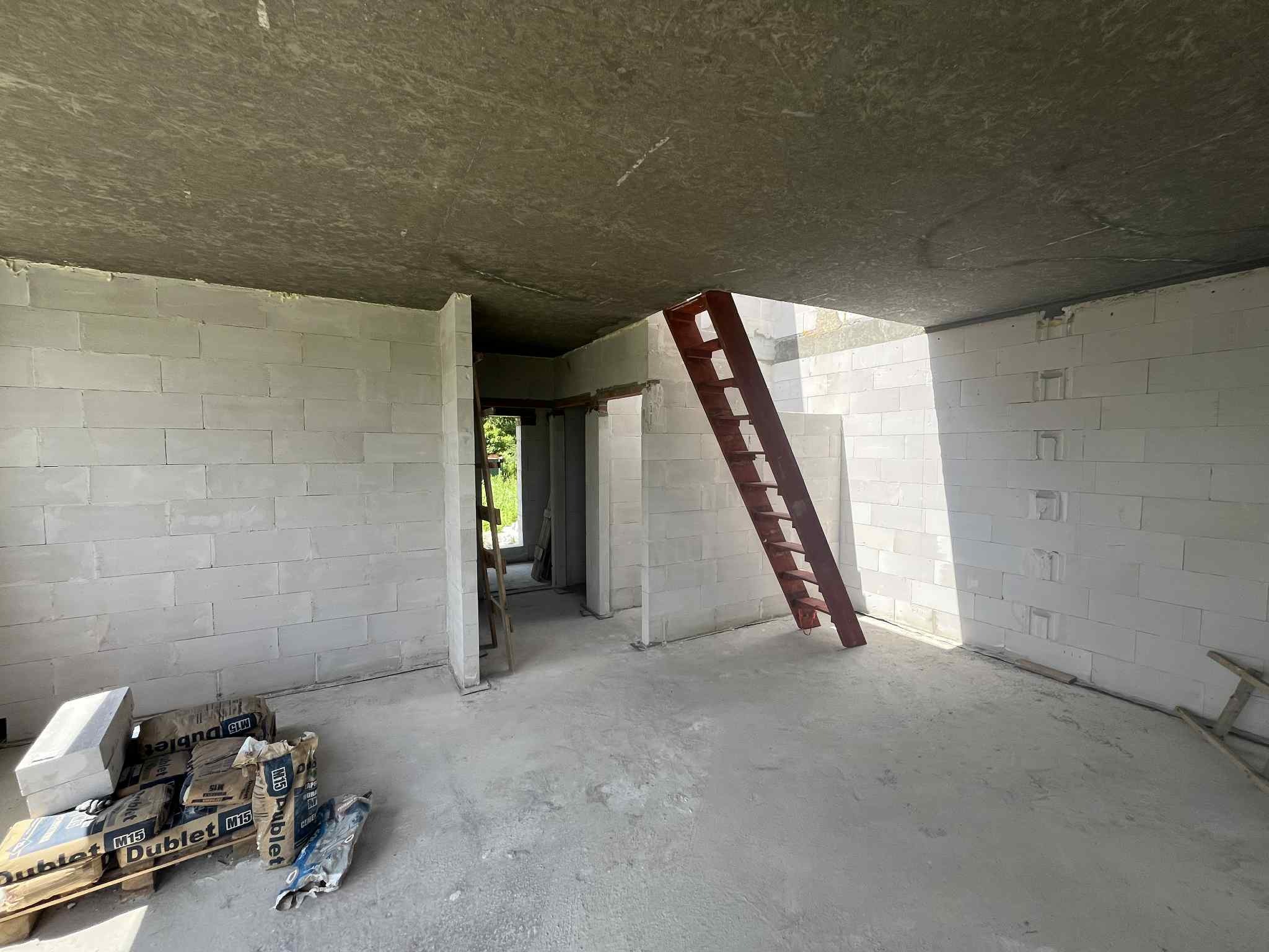 Wnętrze surowego domu w Jeszkowicach: ściany z betonu komórkowego, cement na palecie, drewniana drabina. Widoczny sufit i wylewka podłogowa.