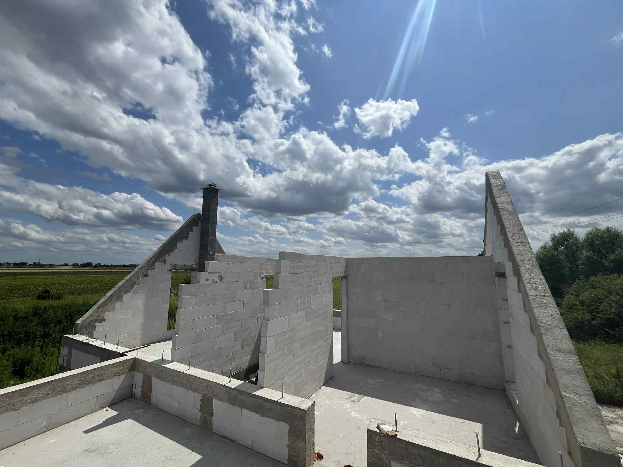 Wznoszenie ścian domu z betonu komórkowego w Jeszkowicach. Widok na konstrukcję w stanie surowym otwartym i słoneczne niebo z chmurami w tle.