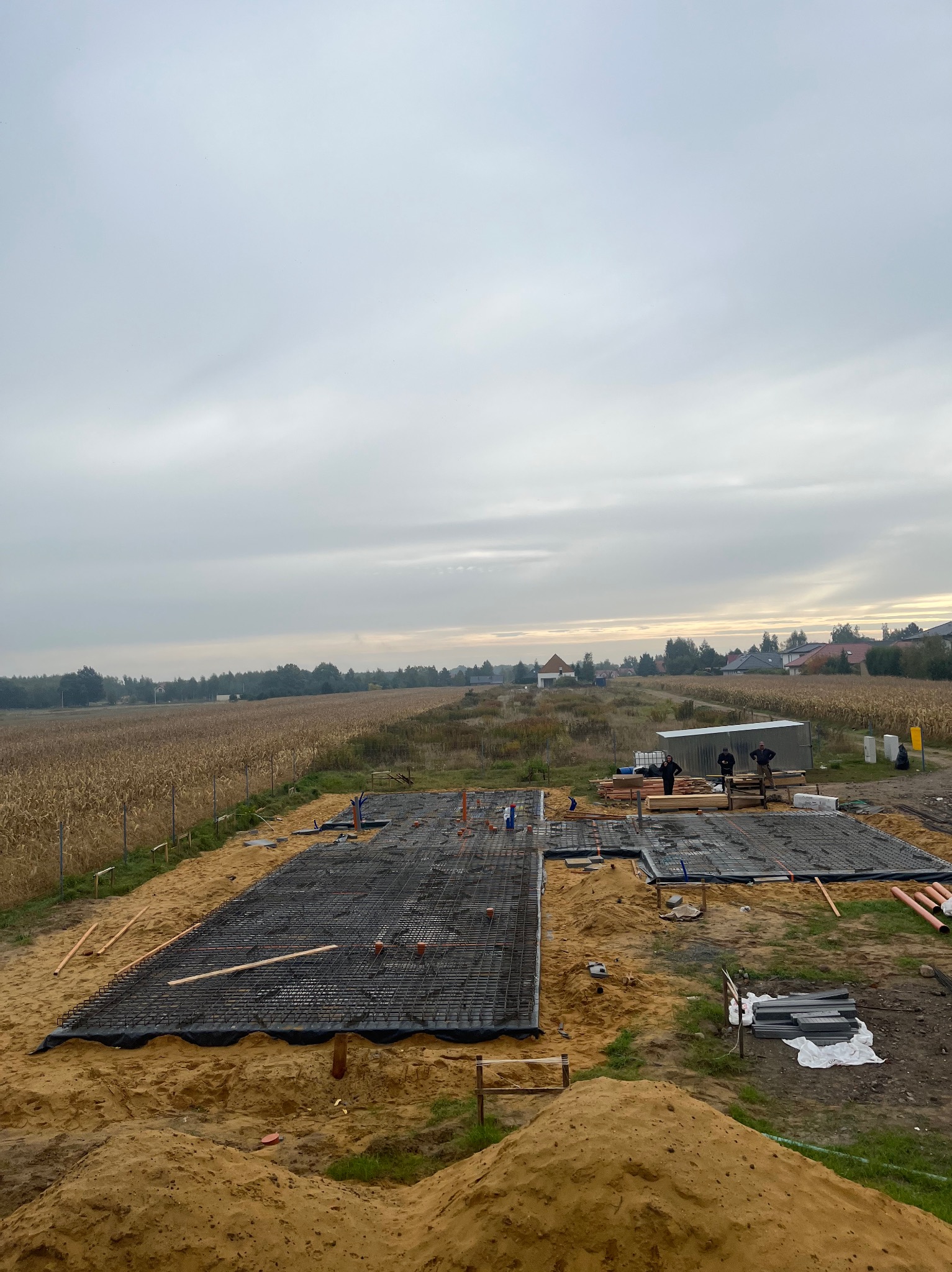 Zbrojenie fundamentów domu jednorodzinnego na tle pola kukurydzy. Widoczne szalunki, pręty zbrojeniowe, folia budowlana i ekipa budowlana przy barakowozie.
