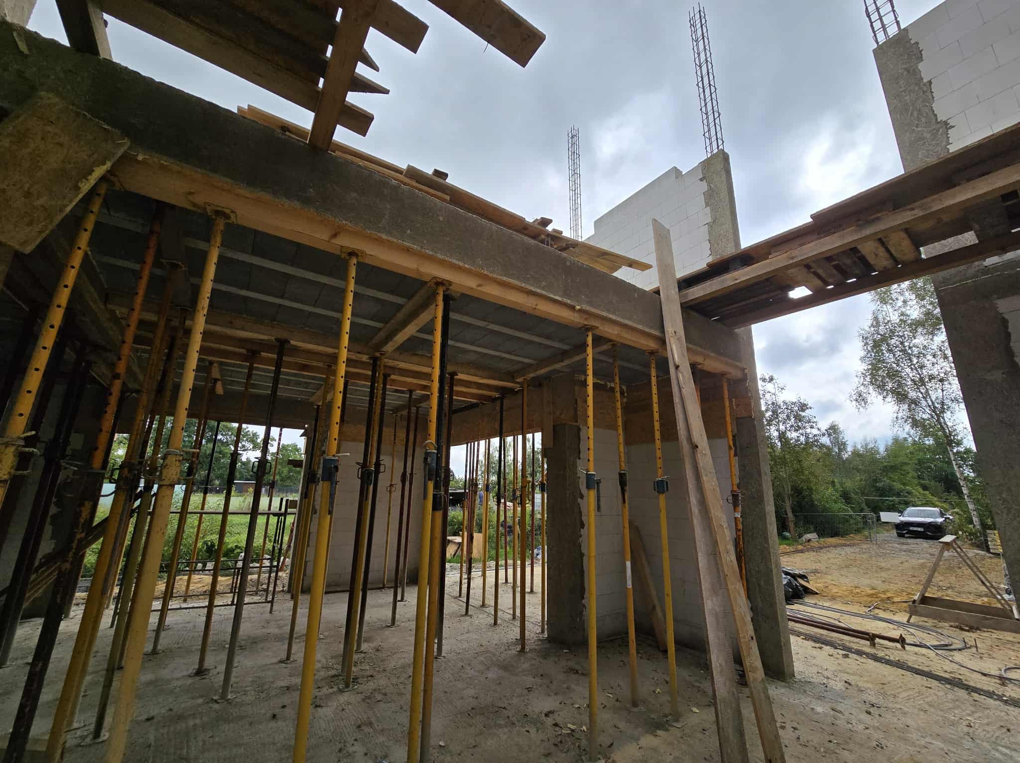 Wnętrze budynku w stanie surowym otwartym. Żółte stemple podtrzymują strop, widoczne ściany z betonu komórkowego i wystające zbrojenia. Na zewnątrz zieleń i samochód.