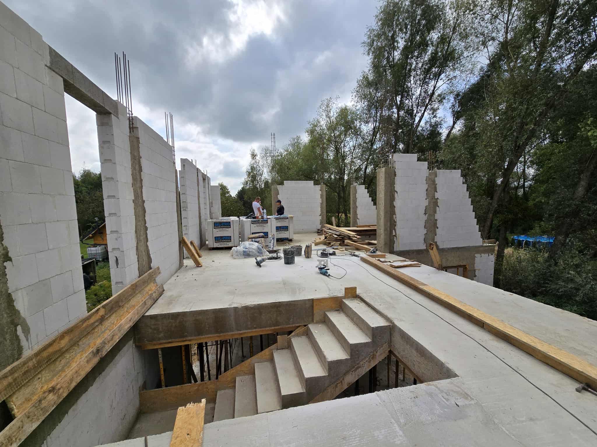 Budowa domu z betonu komórkowego: widok na wznoszone ściany z betonu komórkowego, zbrojenie i betonowe schody w surowym stanie. W tle widoczni pracownicy.