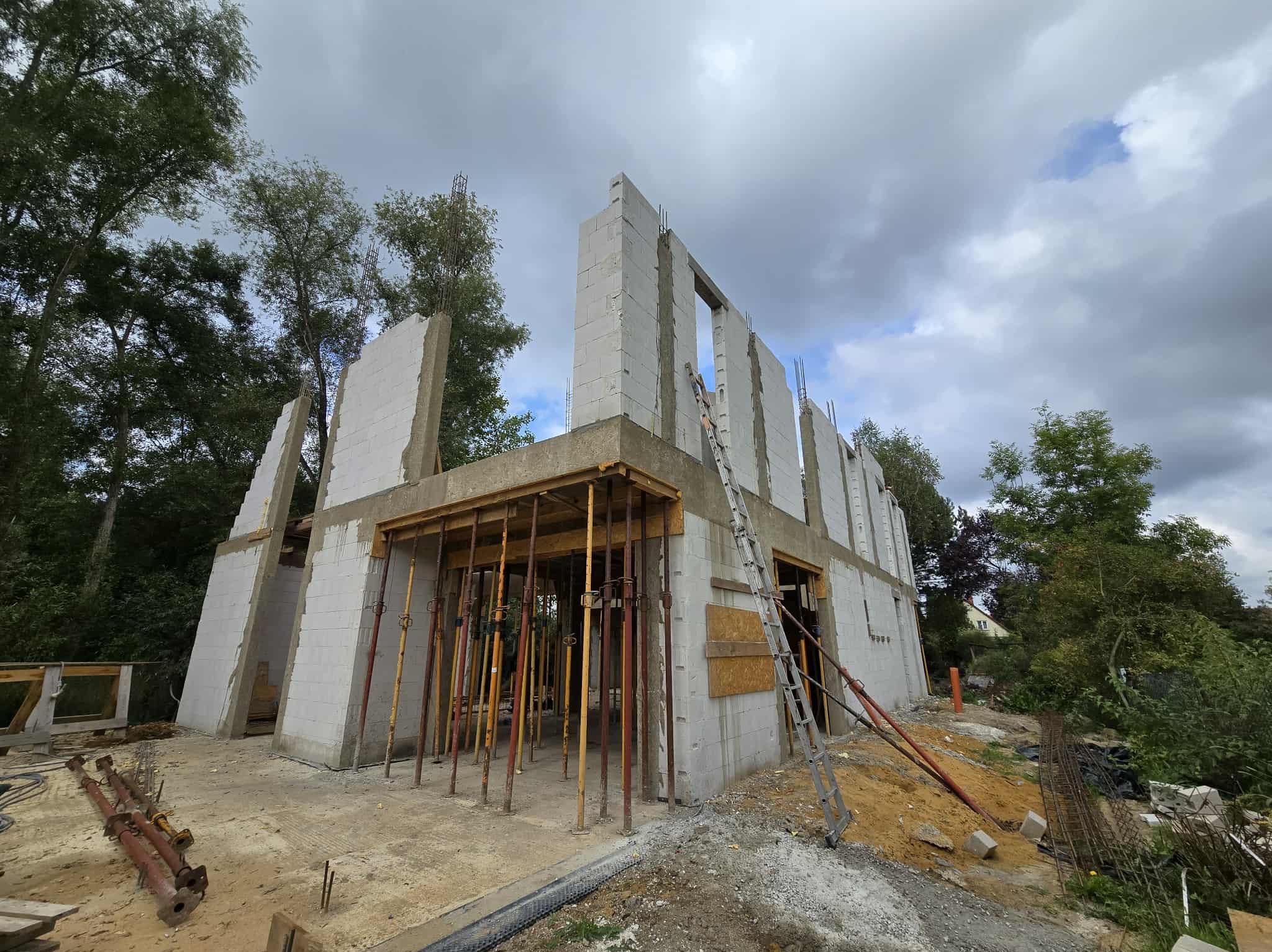 Niedokończony budynek z betonu komórkowego, widoczne podpory szalunkowe i zbrojenie. Częściowo pochmurne niebo w tle, drzewa po bokach.