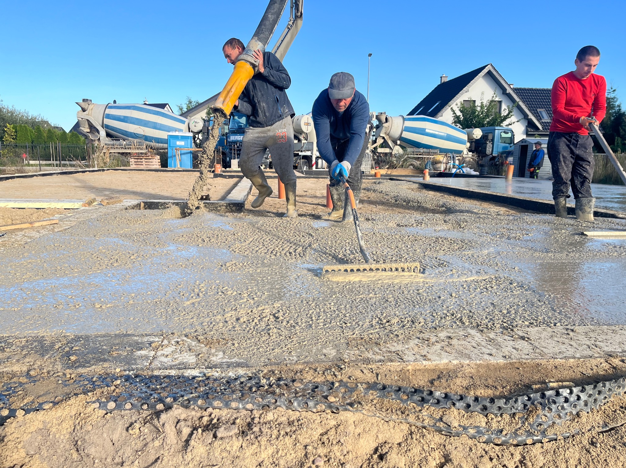 Wylewanie betonu na przygotowany teren przez trzech robotników. W tle betoniarki i dom jednorodzinny z dwuspadowym dachem w słoneczny dzień.