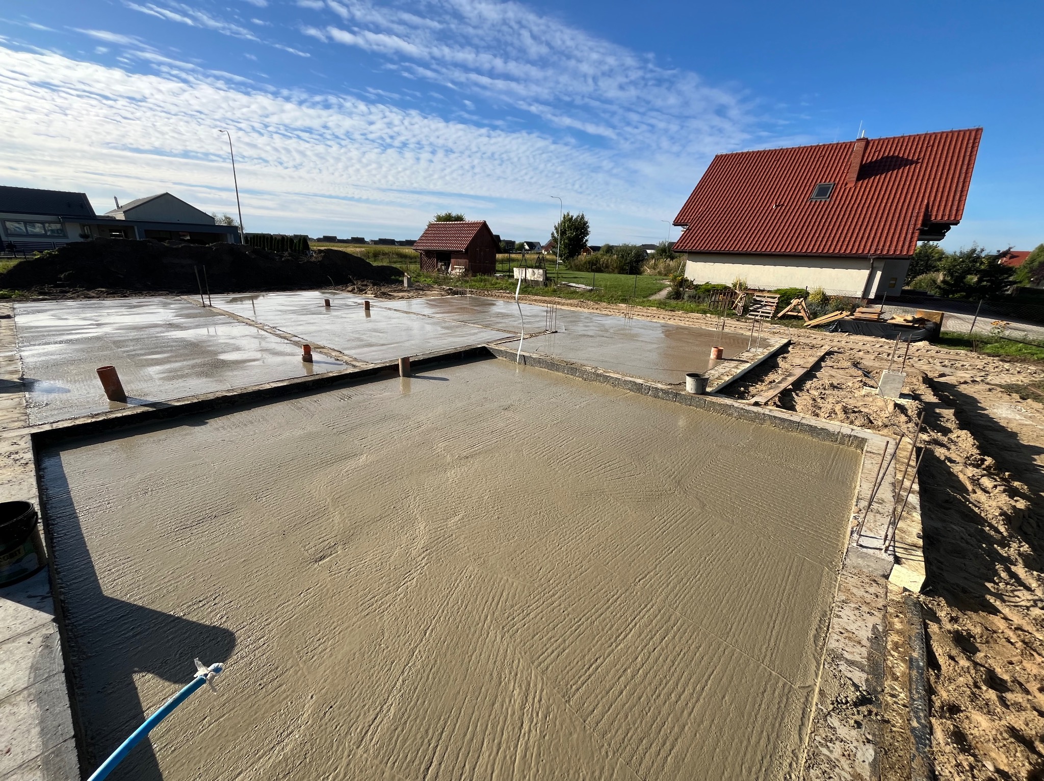 Wylany fundament pod dom jednorodzinny na działce z domami. Widoczne wystające rury i zbrojenie. Błękitne niebo z chmurami w tle. Słoneczny dzień.