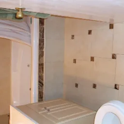 Jasna łazienka z kabiną prysznicową, beżowymi płytkami z brązowymi akcentami, umywalką z szafką i toaletą. Widoczny złoty uchwyt prysznica i zielony ręcznik.