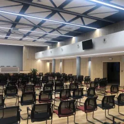 Sala konferencyjna z rzędami czarnych krzeseł z czerwonymi oparciami, drewnianymi panelami ściennymi, sufitem kasetonowym i oświetleniem LED.