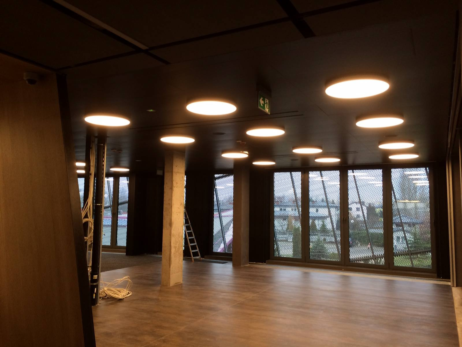 Wnętrze biura w trakcie wykańczania: widoczne okna z siatką, betonowy filar, rozłożone kable i drabina oparta o ścianę, sufit z okrągłymi lampami.