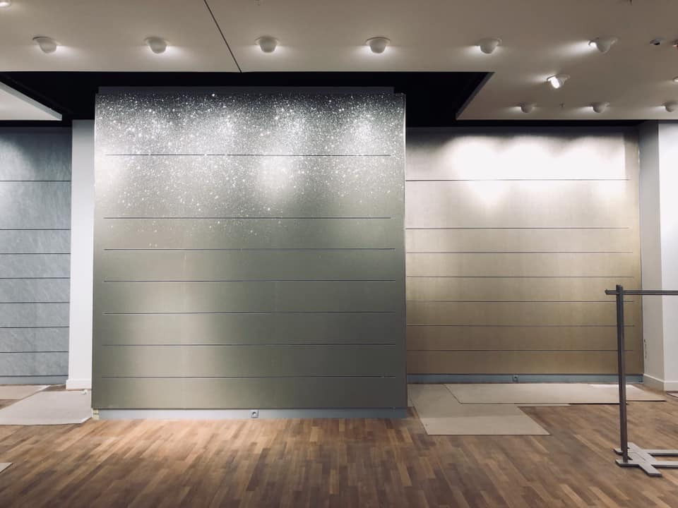 Wykończona ściana z poziomymi panelami w odcieniach szarości i złota, górna część z efektem brokatu, drewniana podłoga w ciepłym odcieniu, widoczne zabezpieczenia podłogi i fragment wieszaka...