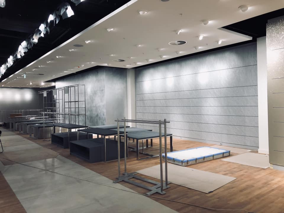 Wnętrze sklepu w trakcie aranżacji: surowe metalowe regały, ciemne stoły ekspozycyjne, zabezpieczone podłogi, jasne oświetlenie sufitowe, szare ściany z poziomymi liniami.