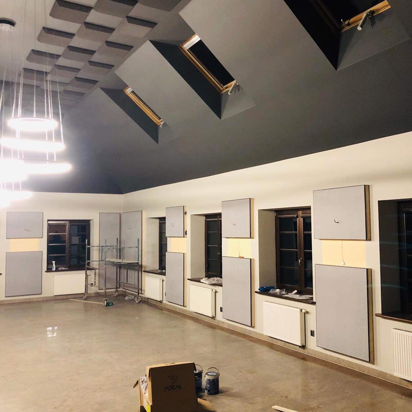 Wnętrze pokoju przed wykończeniem, z zamontowanymi panelami akustycznymi na ścianach, widoczne okna dachowe, sufit w kolorze grafitowym, częściowo pomalowane ściany, puszki z farbą i karton...