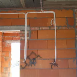 Białe kable elektryczne ułożone na ceglanej ścianie w surowym stanie, widoczne niedokończone ściany z cegieł i betonowy strop. Instalacja elektryczna w trakcie budowy.