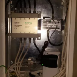 Rozdzielacz antenowy Axing SWE 4-01 w metalowej obudowie z licznymi kablami koncentrycznymi, wzmacniacz AWS-141SE i zasilacz. Widok na instalację wewnątrz skrzynki.