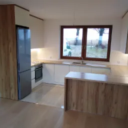 Nowoczesna kuchnia z białymi frontami i drewnianymi akcentami, widok na zewnątrz przez okno z brązową ramą, w tle częściowo widoczne schody.