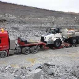 Czerwona ciężar&oacute;wka z naczepą transportuje białą wywrotkę Terex na terenie kamieniołomu, widoczne błoto i kamienie.