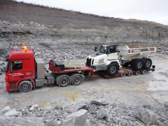 Czerwona ciężarówka z naczepą transportuje białą wywrotkę Terex na terenie kamieniołomu, widoczne błoto i kamienie.