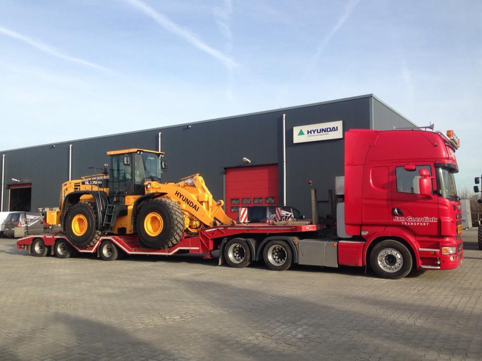 Żółta ładowarka Hyundai HL770-9A transportowana na czerwonej platformie niskopodwoziowej ciągniętej przez czerwoną ciężarówkę Jan Geerdink Transport na tle szarego budynku z logo Hyundai.