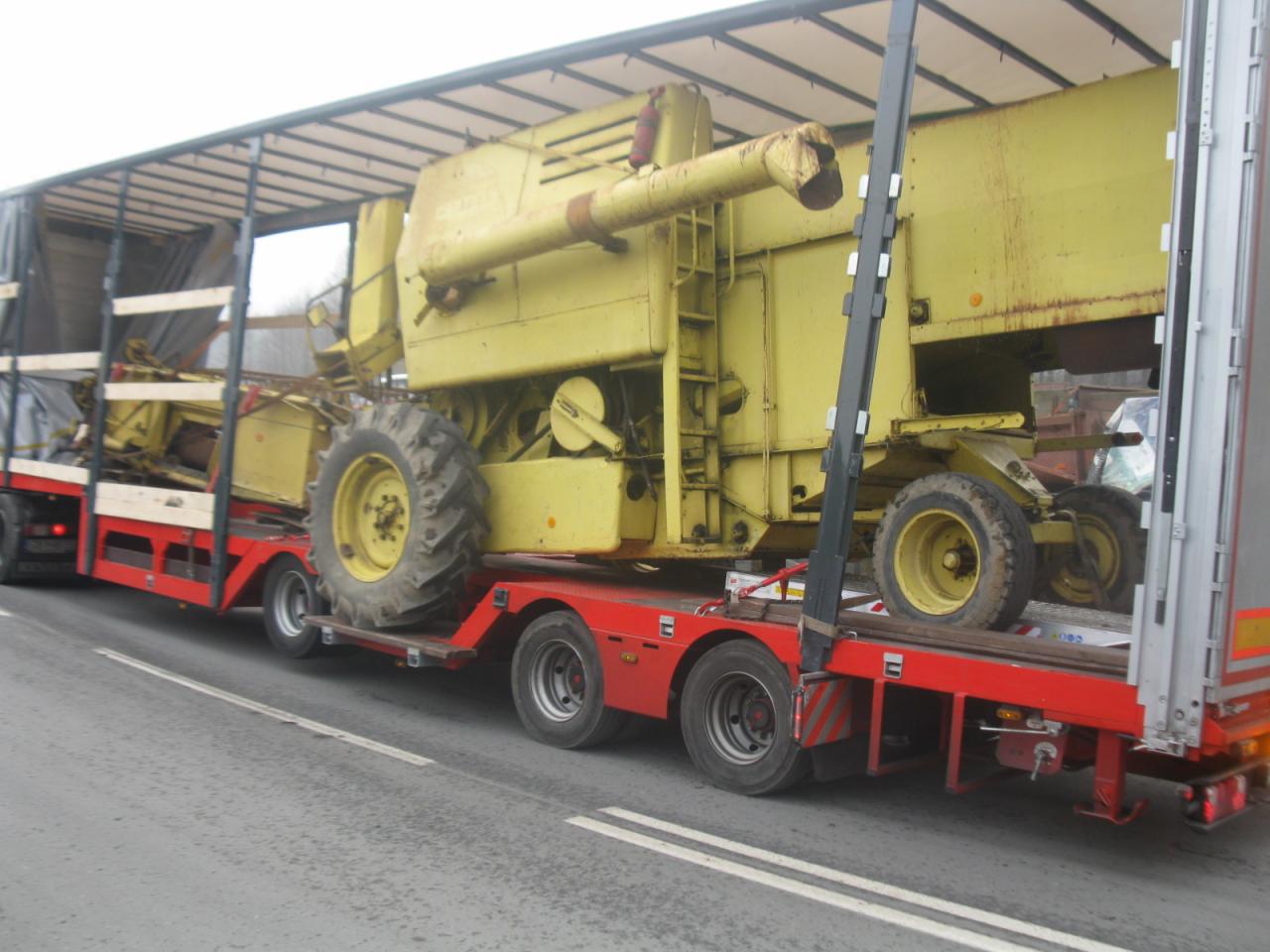 Żółty kombajn rolniczy przewożony na czerwonej platformie niskopodwoziowej pod plandeką ciężarówki, widok z boku podczas jazdy.