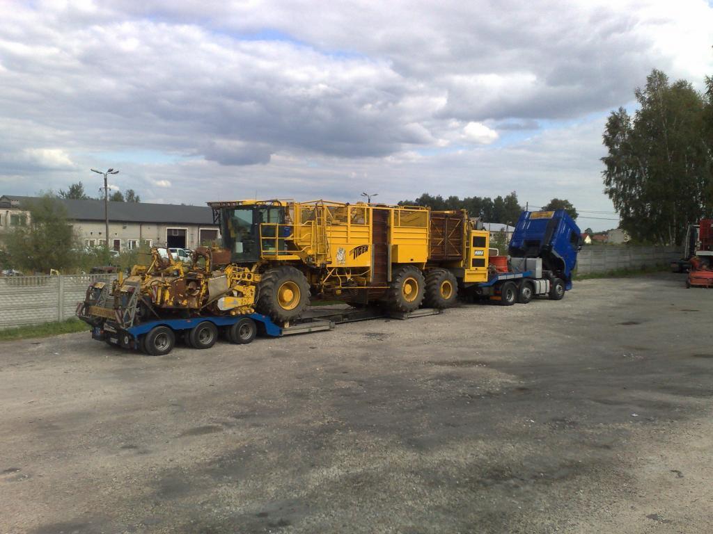 Niskopodwoziowa platforma z żółtym kombajnem rolniczym na lawecie, transportowana przez niebieski ciągnik siodłowy w Warszawie.