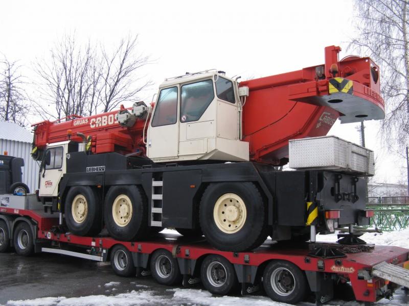 Czerwono-biały dźwig samojezdny marki Liebherr przewożony na niskopodwoziowej platformie transportowej, widok z boku, teren częściowo pokryty śniegiem.