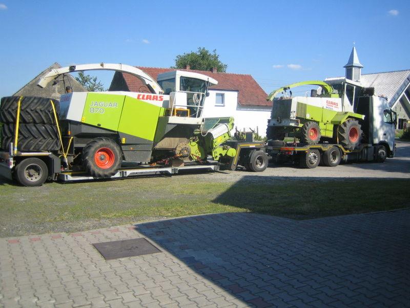 Ciężarówka z naczepą przewozi dwa kombajny rolnicze marki Claas Jaguar 870, na tle wiejskiego krajobrazu z domami i kościołem.