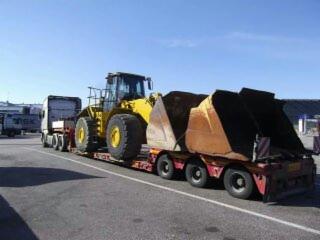 Żółta ładowarka na niskopodwoziowej platformie transportowej, ciągnięta przez biały ciągnik siodłowy. Widoczne metalowe łyżki do załadunku. Tło to parking i budynki.
