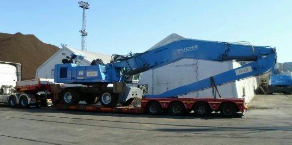 Niskopodwoziowa platforma transportowa przewozi niebieską koparkę Fuchs na tle magazynu i hałdy materiałów.